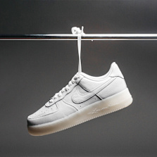 A white low‑top sneaker with a visible Nike swoosh hangs from a metal rod by a white zip tie against a dark gray background.