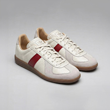 Pair of off‑white low‑top sneakers with a red side stripe, suede panels, beige laces, and a gum rubber sole, photographed on a light gray background.