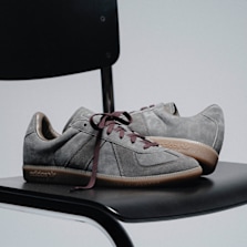 Pair of adidas BW Army sneakers in grey suede with gum soles and burgundy laces, displayed on a black chair in a minimalist studio setting.