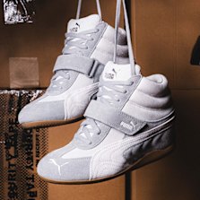 White Puma Wmns Speedcat Wedge sneakers suspended in front of stacked cardboard boxes, highlighting the elevated wedge sole, suede overlays, and sporty motorsport-inspired design.