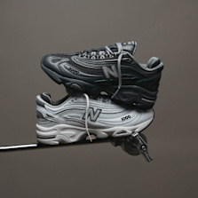 Two pairs of New Balance 1000 sneakers in monochrome tones—one black, one white—balanced on a metallic rod against a dark background.