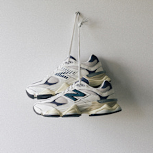 A pair of New Balance 9060 sneakers in beige and navy, hanging by their laces against a plain white wall.