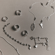 "A collection of silver rings and necklaces laid out on a neutral background, featuring star-shaped designs, keys, a four-leaf clover, a dice, and other small charms."
