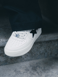 Close-up of a white sneaker with a black star design, worn by a person standing on a concrete step. The shoe features perforations on the toe box, white laces with a metallic lace lock, and is partially covered by dark pants.
