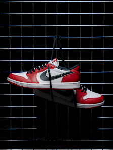 A pair of Nike Air Jordan 1 Low sneakers in red, white, and black, hanging by their laces against a black wire grid background.