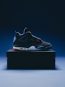 The image showcases the Air Jordan 4 "Rare Air" in a striking design, featuring a dark colorway with black suede and leather accents. The shoe rests on its original box, highlighted by subtle red and blue details on the midsole and tongue. The lighting emphasizes the sleek contours and iconic elements of the Air Jordan 4, giving it a bold, premium appearance.