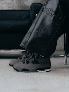 Close-up side view of a person wearing dark grey Air Jordan 4 sneakers with black mesh detailing, standing on a light carpet. The person is dressed in wide brown leather pants with embroidered floral and geometric patterns. A black leather sofa is partially visible in the background.