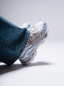 A person is seen from the ankle down, wearing light-wash jeans and a white and light blue ASICS Gel-NYC sneaker with tan accents. The image is taken from a low angle, and the person's foot is propped up, revealing the detailed design of the shoe's side and sole. The background is a plain, bright white.
