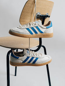 A pair of cream-colored Adidas sneakers with blue stripes and a gum sole, suspended by laces in front of a light wooden chair.