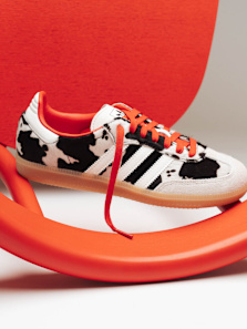 A sneaker with a black-and-white cow print pattern, three black stripes on the side, bright orange laces, and an orange inner lining. The shoe has a beige gum sole and is displayed on a curved orange chair against a neutral background.