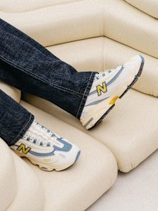 Person wearing New Balance 1000 sneakers in a white, blue, and yellow colorway, paired with dark denim jeans, sitting on a cream-colored, modern cushioned seat.