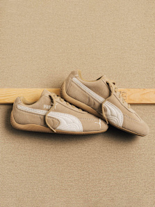 A pair of tan Puma Speedcat sneakers with cream-colored side stripes and gum soles, resting on a wooden ledge against a light brown background.