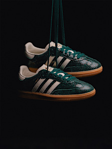 A pair of Adidas Samba sneakers, with a dark green, textured, scale-like upper and a classic gum sole, hangs against a completely black background. They are suspended by their laces, which are also dark green. The iconic white Adidas three stripes are visible on the side of each shoe.