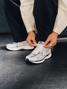 A person tying the laces of a silver and white Nike Zoom Vomero sneaker, seated on a dark floor while wearing black pants and a light jacket.