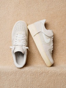 Neutral grey Nike Air Force 1 sneakers with suede uppers, shown as a pair from top and side view on a beige textured background.
