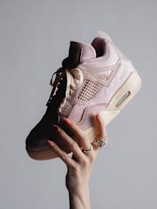 Hand holding an Air Jordan 4 Retro ‘Iced Carmine’ sneaker, shown from a side angle with soft pink suede upper, mesh panels, translucent lace wings, and cream‑colored midsole with visible Air cushioning, photographed against a clean grey studio background.