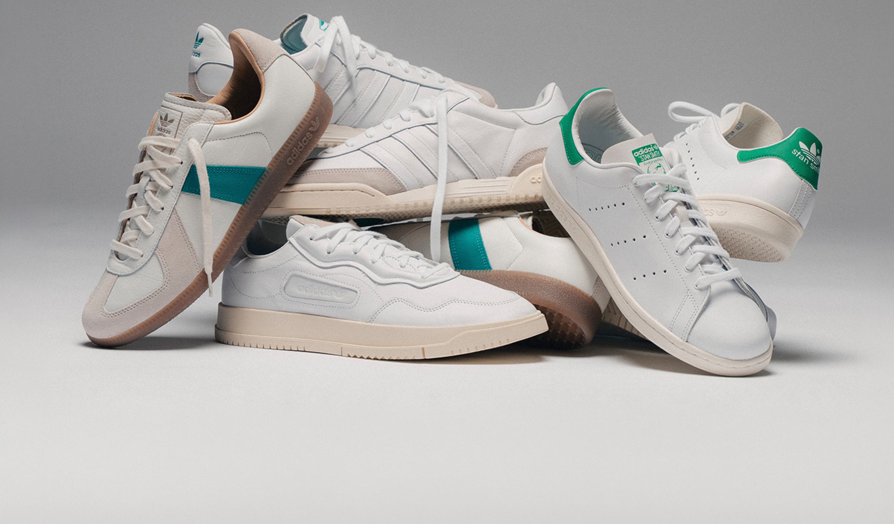 Adidas Court Pack sneakers in white leather with green accents, including multiple classic silhouettes arranged in a stacked display against a clean studio background. Geben Sie Ihr Feedback zu BizChat ab.