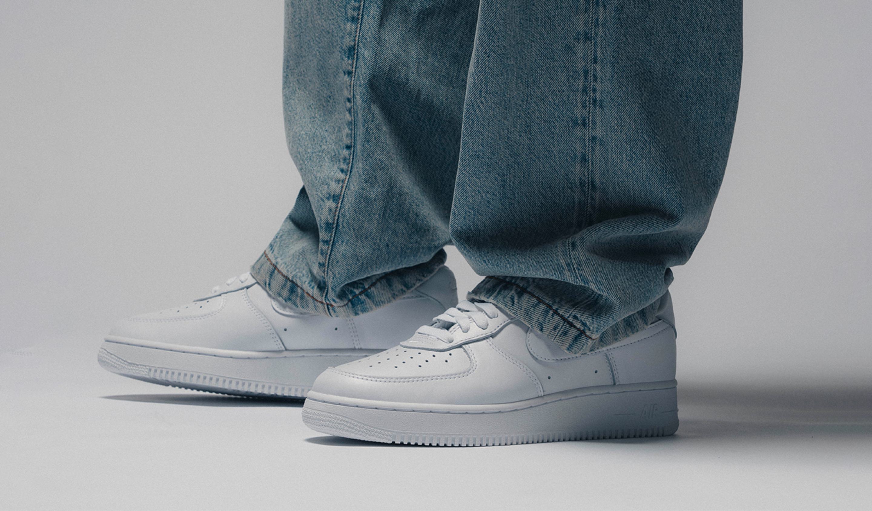 Close-up of the Nike Air Force 1 “Triple White” (2001 Shape) in all-white leather, worn with loose blue denim jeans, highlighting the classic low-top silhouette and clean minimalist design.