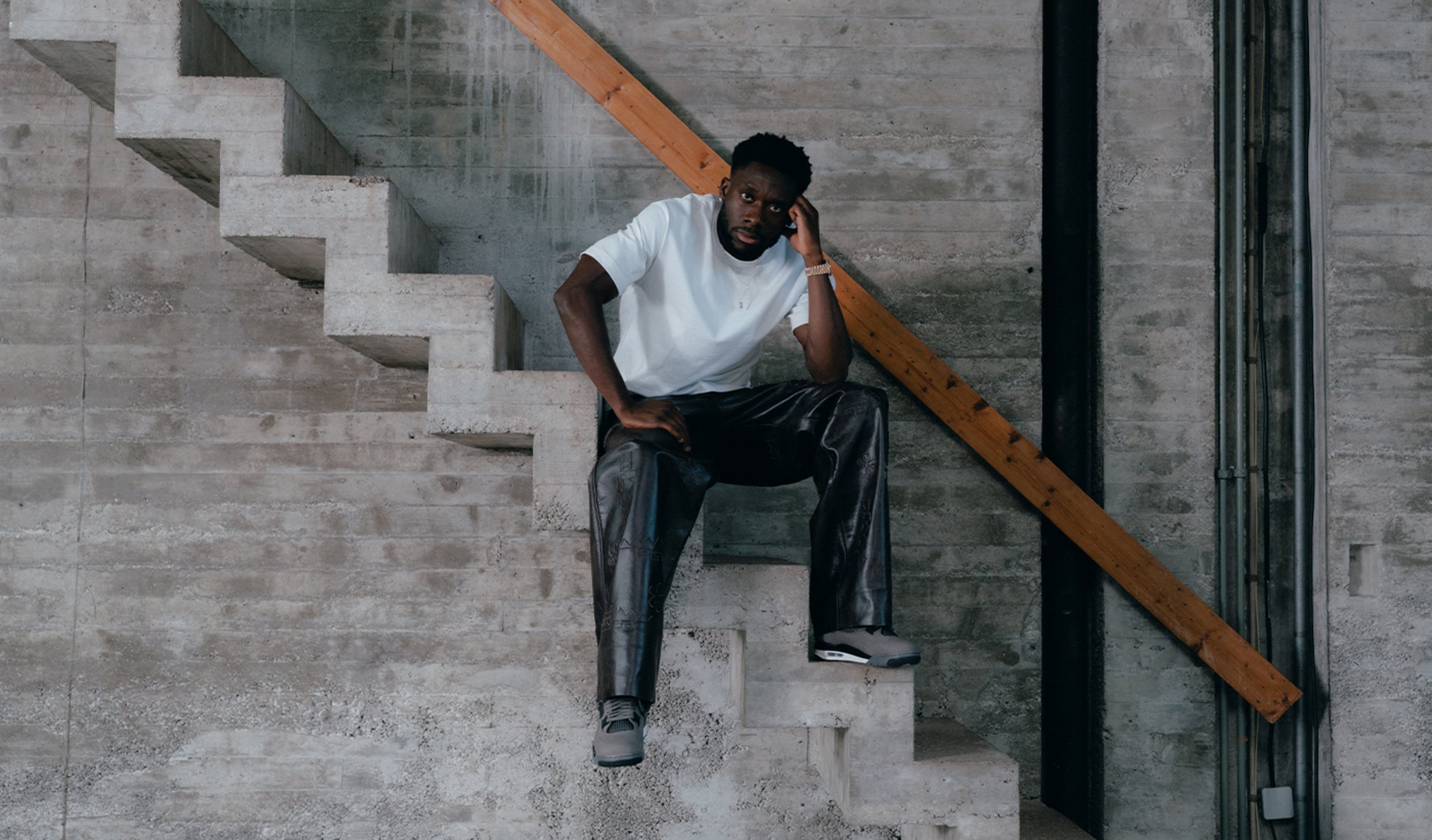 A man wearing a white t-shirt, black leather pants with subtle embossed detailing, and grey Air Jordan 4 Cavestone sneakers, sitting on a concrete staircase in an industrial setting.