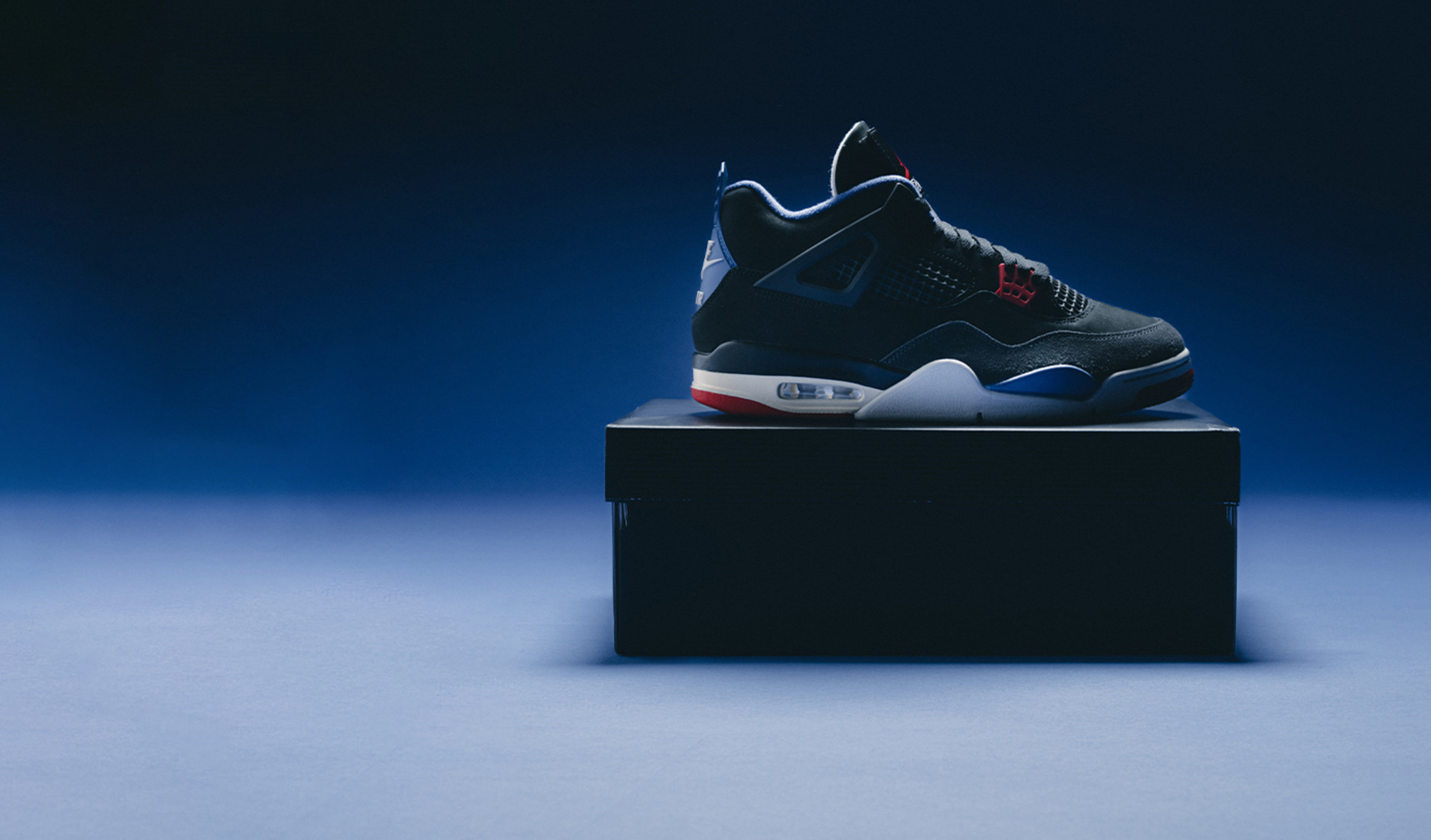 A single Air Jordan 4 sneaker in black, red, white, and blue colorway displayed on top of a closed black shoebox, set against a dark blue gradient background with dramatic lighting.