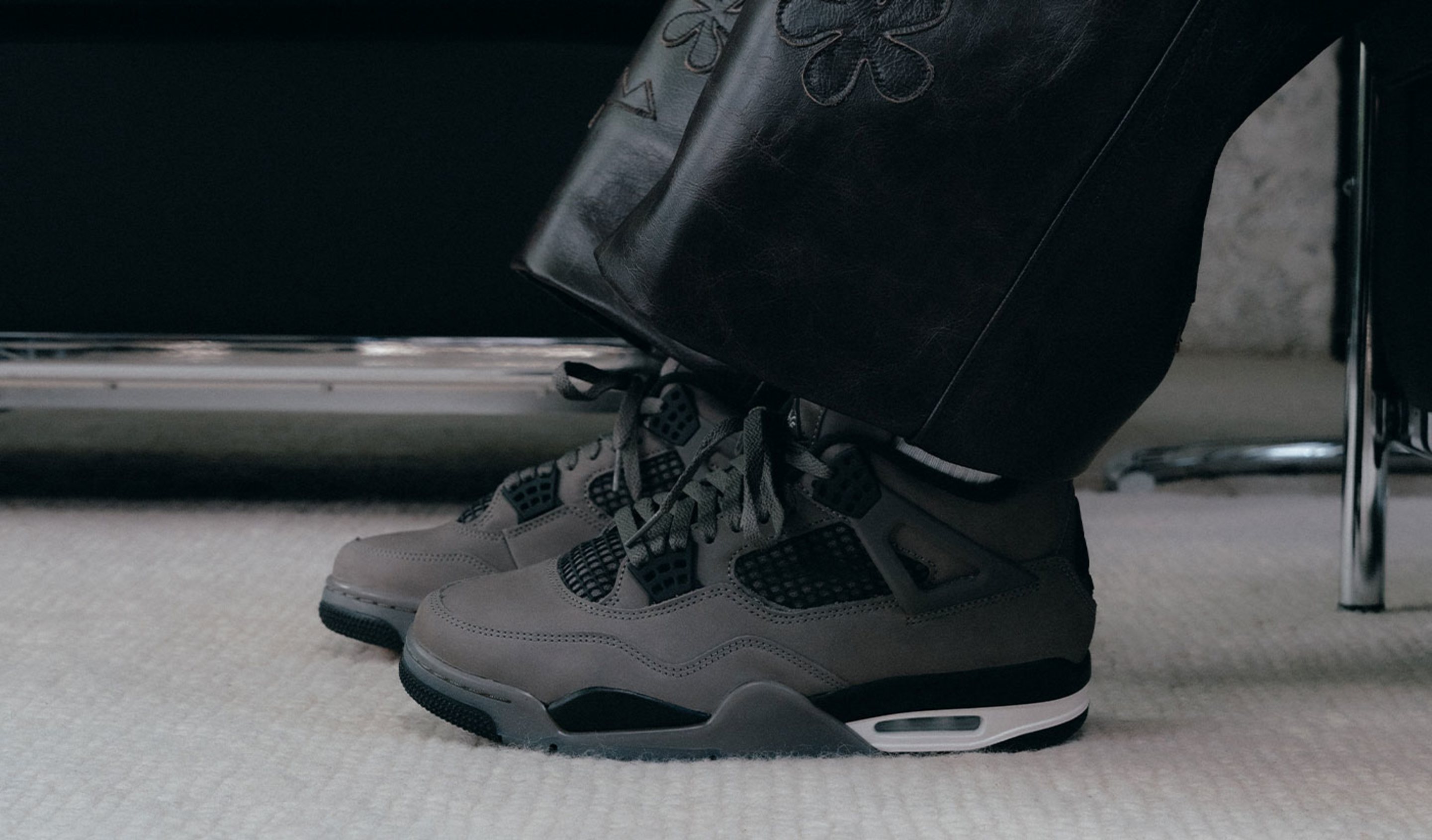 "A close-up side view of someone wearing dark grey Air Jordan 4 sneakers with black and white accents, paired with black leather pants with floral embroidery."