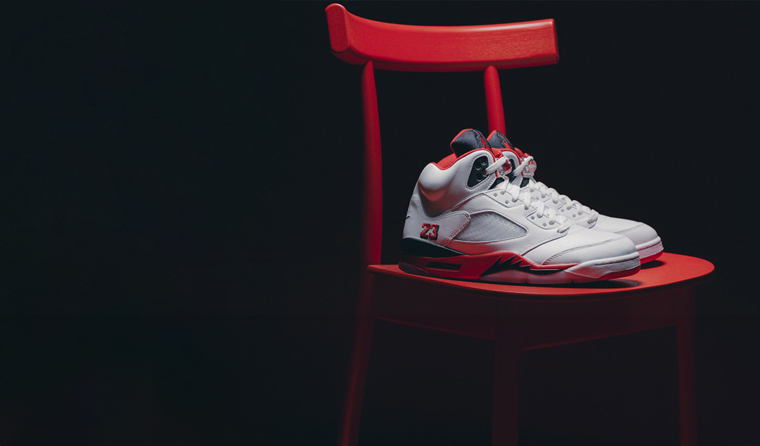 A pair of white and red Air Jordan 5 sneakers placed on a bright red chair. The sneakers feature a red inner lining, black and red midsole with a flame design, and the number 23 stitched on the side. The background is dark, creating a dramatic contrast with the chair and shoes.