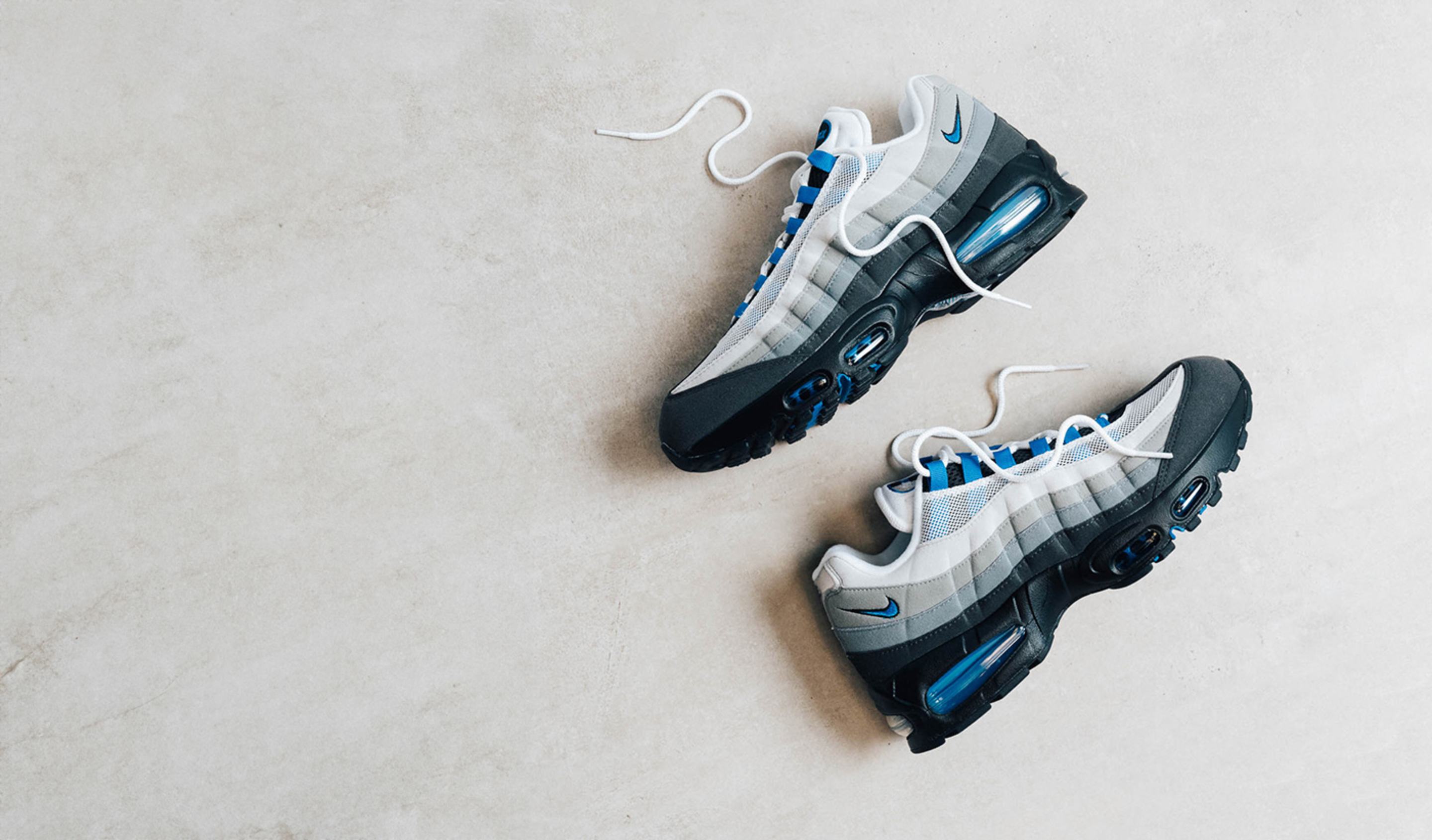 A pair of Nike Air Max 95 sneakers in gradient grey with black soles, blue accents, and visible Air units, placed on a light beige surface with untied white laces.