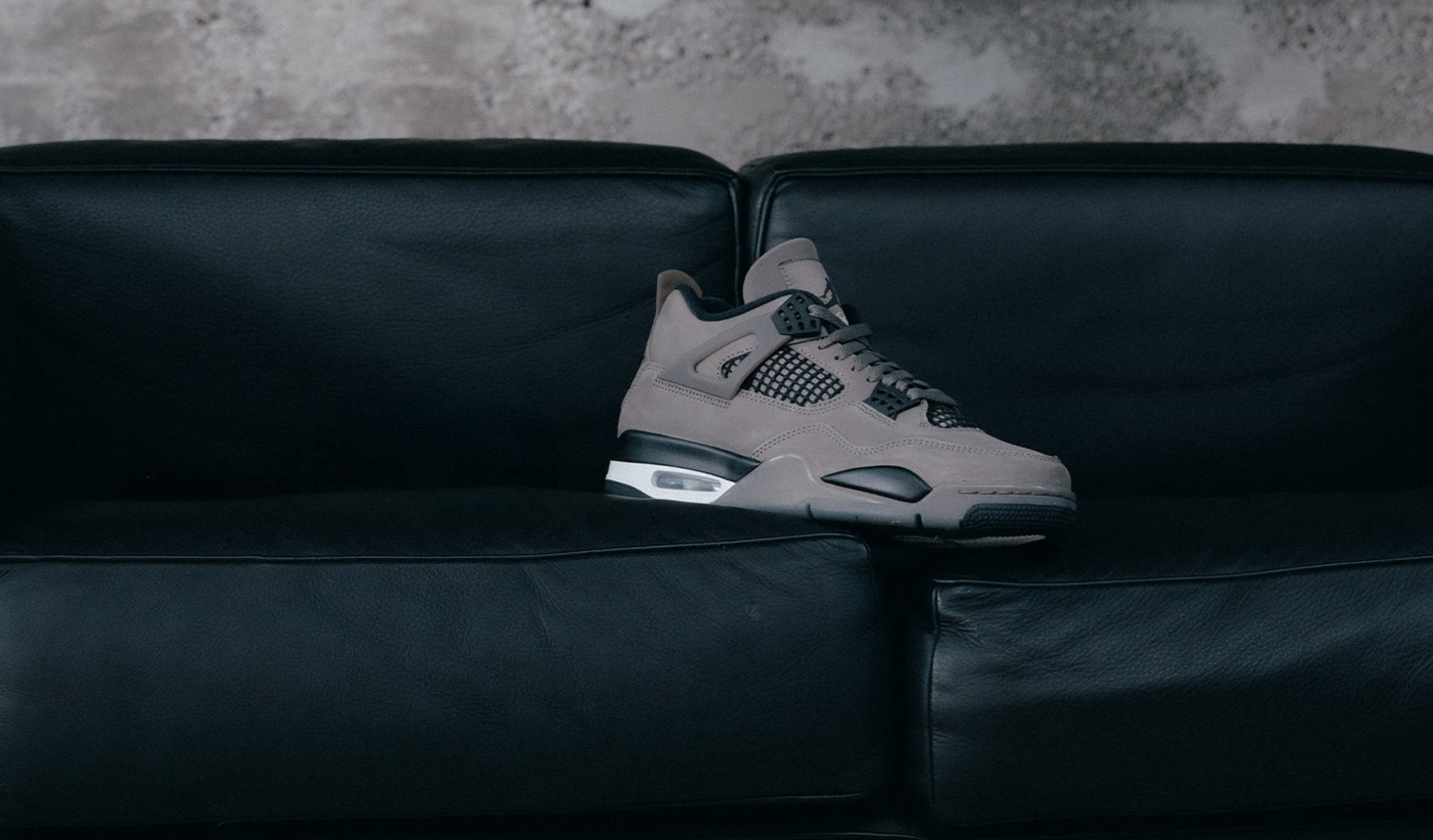 A single taupe-grey sneaker is placed on a black leather couch, set against a rough concrete wall. The shoe resembles an Air Jordan 4 with visible Air cushioning, black and white accents, and mesh side panels.