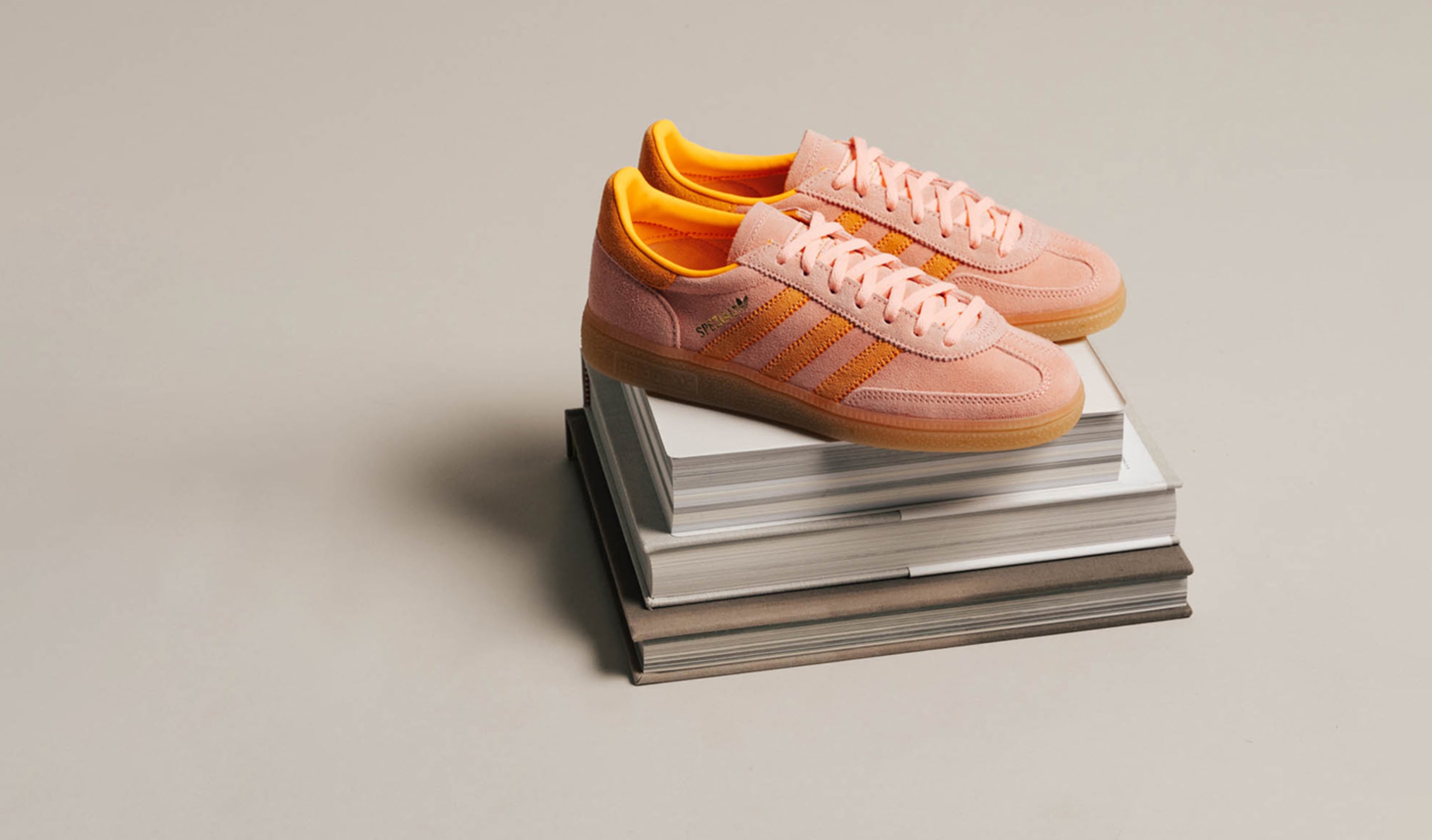 A pair of Adidas Handball Spezial sneakers in soft pink suede with bright orange stripes and inner lining, placed on top of a stack of books against a neutral beige background.