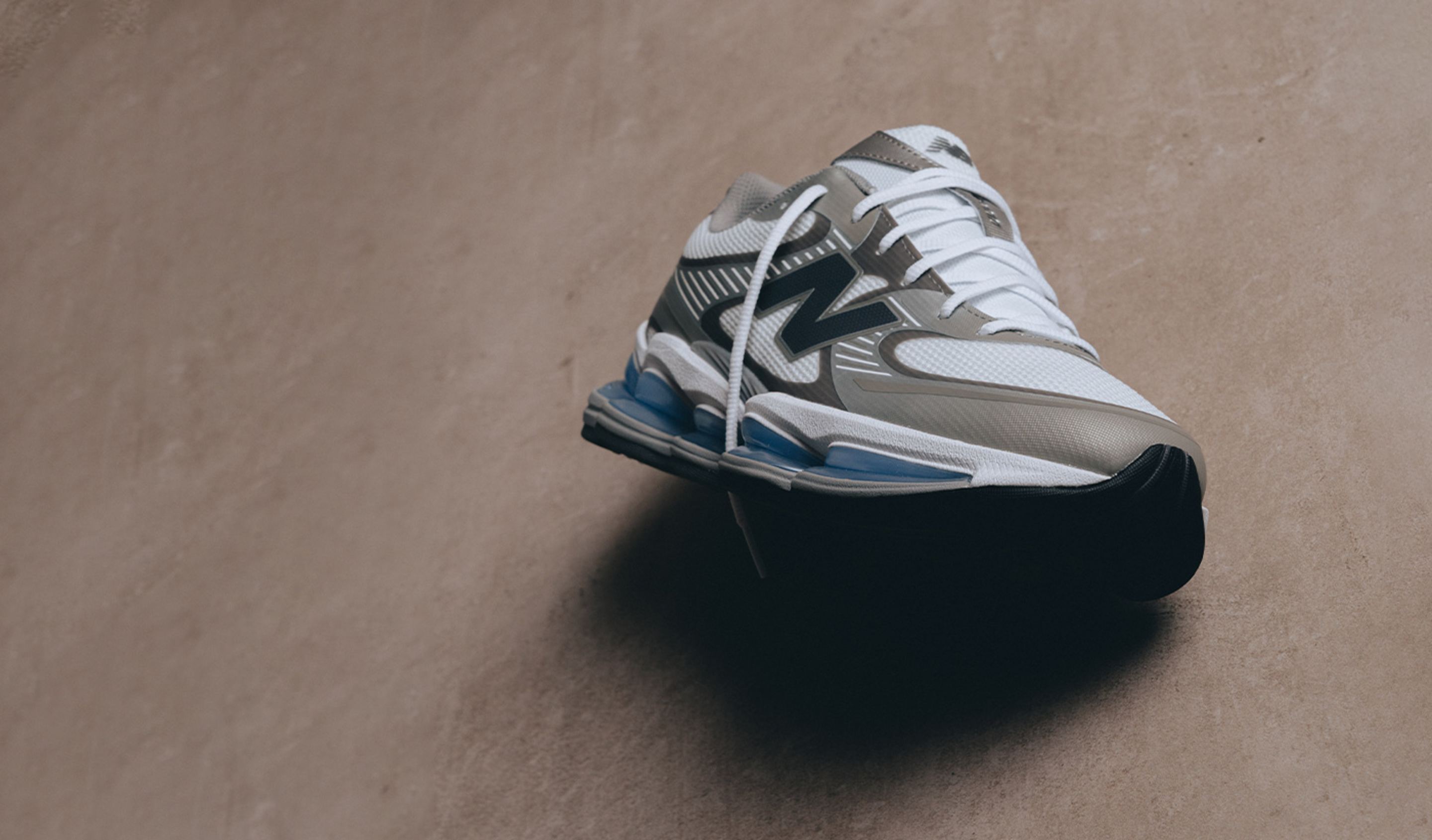 A single New Balance sneaker is showcased on a neutral-toned background, featuring white mesh, grey overlays, and blue cushioning details.