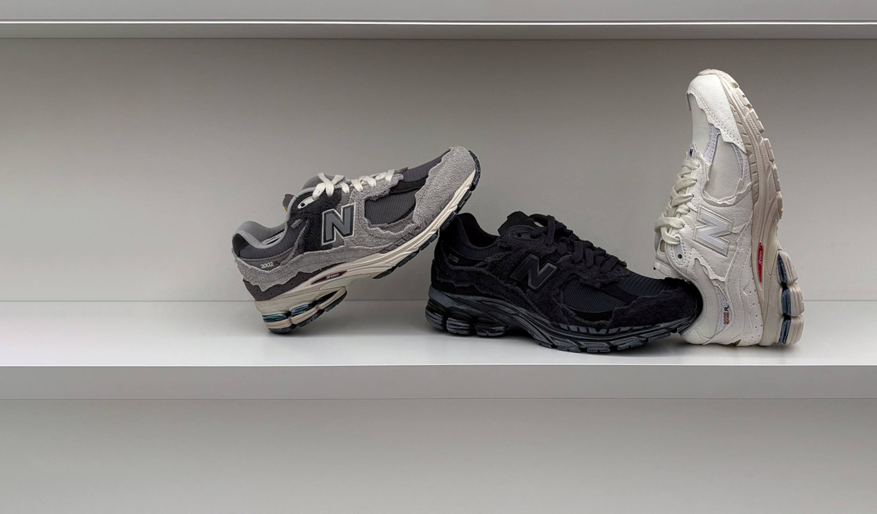 Three New Balance 2002R sneakers displayed on a minimalist white shelf: one in grey with a deconstructed suede look, one in black with rugged suede overlays, and one in off-white with distressed detailing and red accents.