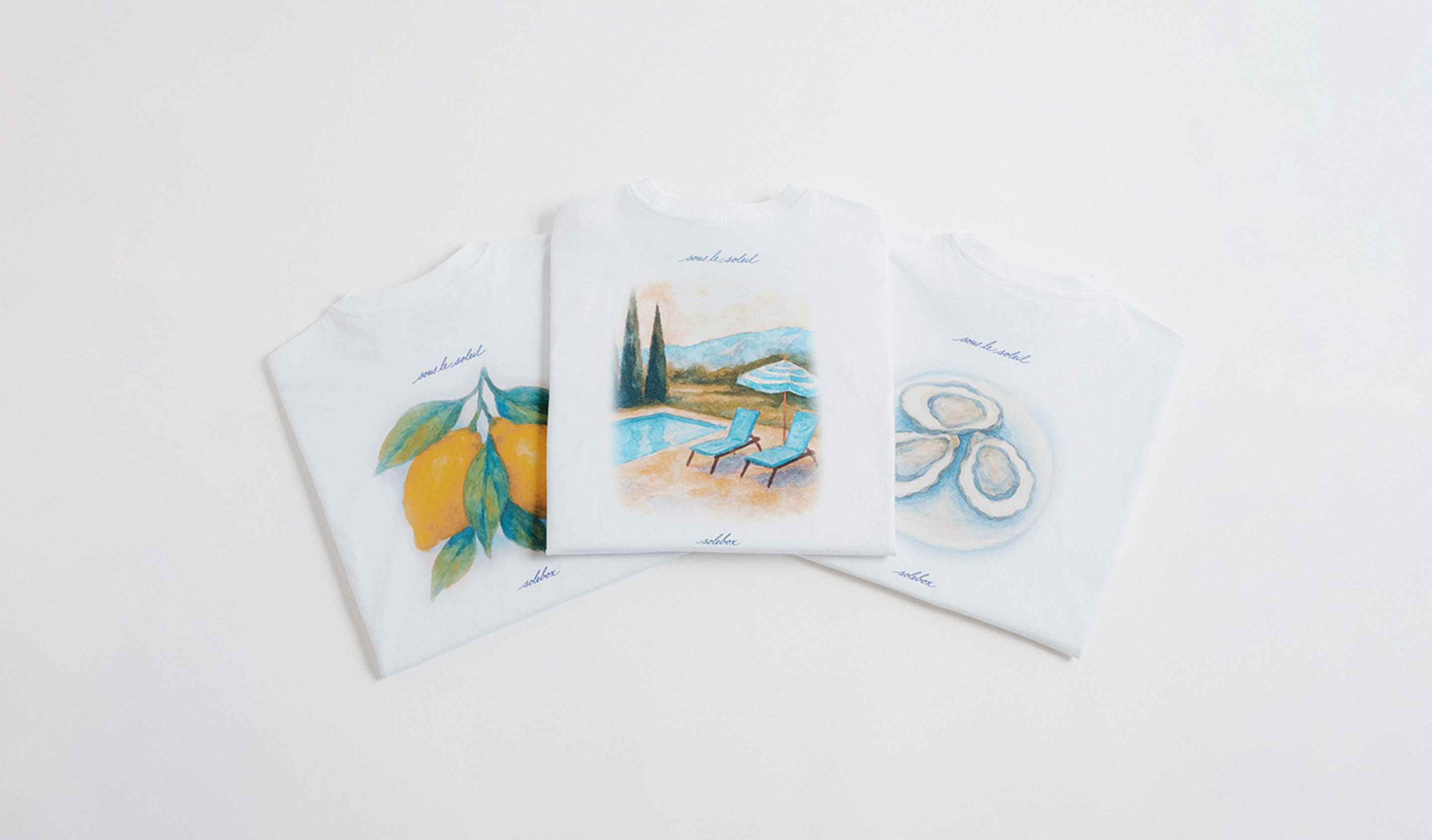 Three folded white T-shirts laid out side by side, each featuring a colorful illustration: lemons on the left, a poolside scene with chairs and umbrella in the center, and oysters on the right.