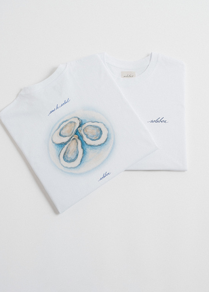 "Two folded white T-shirts, one featuring an oyster illustration with the words 'sous le soleil' and 'solebox', and the other with a small 'solebox' logo on the chest."