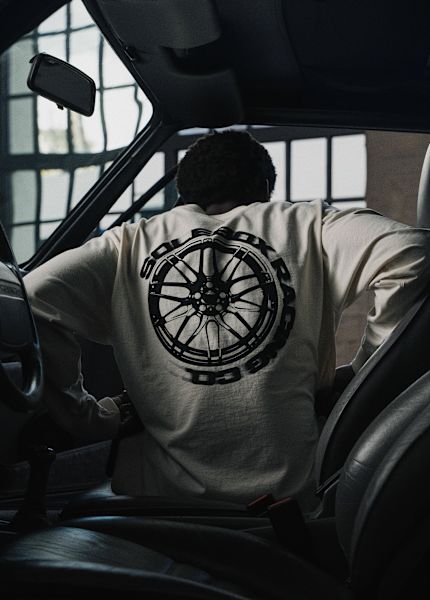 Person seen from behind wearing a cream long-sleeve shirt with a large solebox racing wheel graphic on the back, holding a jacket over the shoulder.