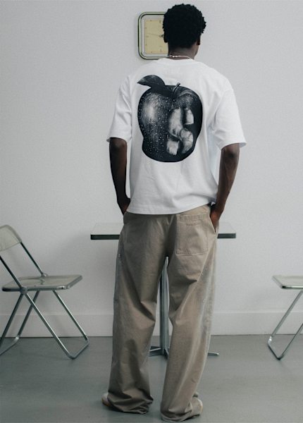 Back view of a person wearing the solebox Apple Tea T-shirt with a large apple graphic print, paired with loose beige trousers in a minimalist interior. Geben Sie Ihr Feedback zu BizChat ab.