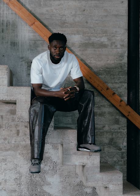 A man sits on a concrete staircase against a rough concrete wall with a diagonal wooden beam. He is wearing a plain white t-shirt, black leather pants with subtle patterns, and gray sneakers. He looks directly at the camera with his hands clasped.