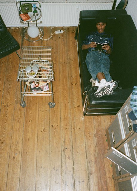 A man is lying on a black leather sofa in a cozy room with wooden floors, reading a book or tablet. He is wearing a blue satin shirt, jeans, white socks, and black-and-white sneakers. Next to him is a small metal coffee table on wheels, holding cups, snacks, and magazines. A black lounge chair is nearby, and there is a side table with a vintage telephone, books, and a lamp. Filing cabinets are partially visible in the bottom right corner. The scene is viewed from above.
