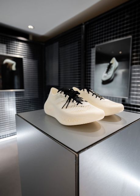 A pair of modern, cream-colored clima cool laced sneaker with a futuristic, lattice-like design and black laces displayed on a sleek pedestal in a minimalistic showroom.