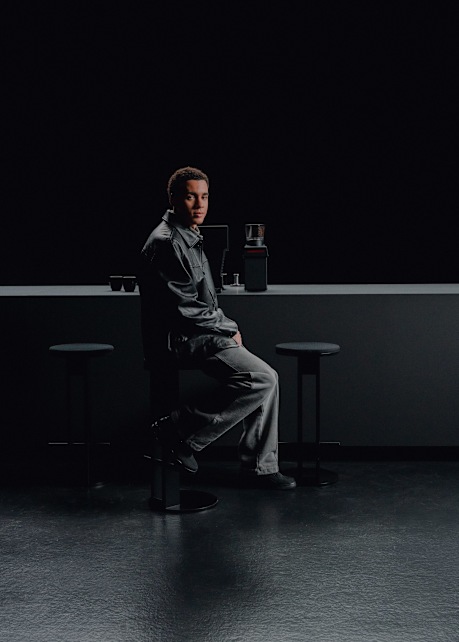 A person seated on a tall stool in a minimalist, dark indoor setting. The individual is wearing a loose-fitting dark outfit, and there are two additional stools and a sleek counter behind them with a coffee grinder and cups placed on top.