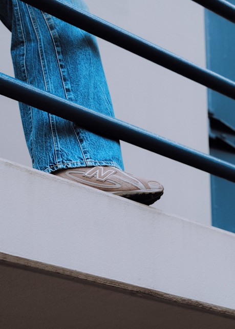 Close-up of a person wearing beige New Balance 204L sneakers with wide-legged blue jeans, standing on a concrete balcony with a metal railing.