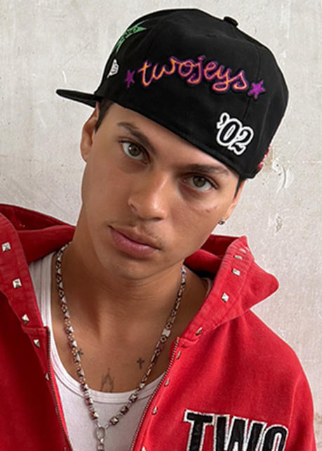 Person wearing a black “twojeys” cap with colorful lettering and a red studded jacket with a “TWO” patch.