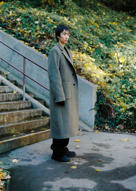 A person standing outdoors near a concrete staircase with metal railings, wearing a long gray overcoat and black pants. The background shows a sloped area covered with green foliage and scattered yellow leaves.
