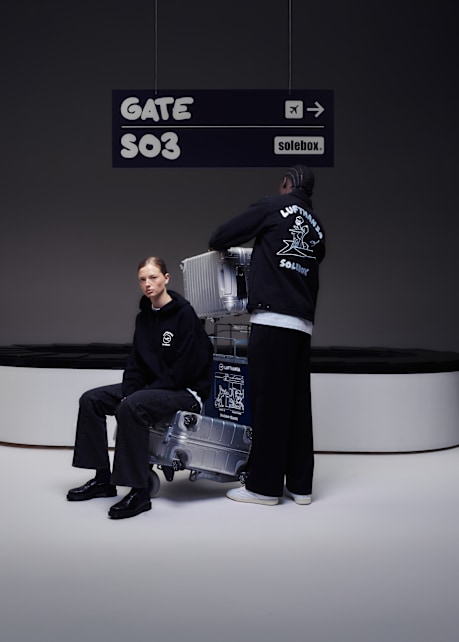 Two individuals dressed in black streetwear are positioned in front of a modern airport-themed backdrop. One person is seated on a silver suitcase, while the other stands beside it, leaning on another suitcase. Both wear oversized black jackets featuring bold white graphic designs on the back. Above them hangs a dark sign reading “GATE S03” with an arrow and the word “solebox.” The setting is minimalistic with a circular white platform and a dark background, creating a sleek, travel-inspired aesthetic.