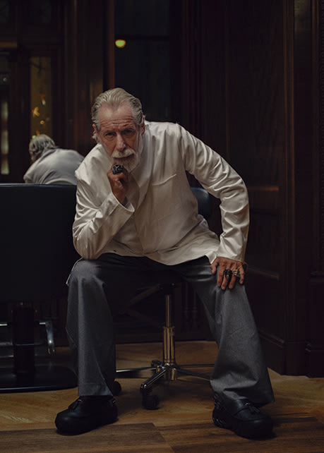 Person seated on a salon chair wearing a white button-up shirt, wide-leg grey trousers, and black Crocs Echo clogs in a dark wood-paneled interior.