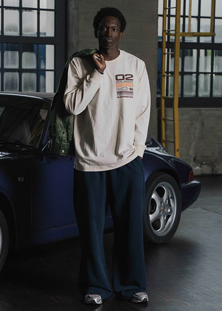 Person wearing a cream long-sleeve graphic shirt and dark trousers, standing in front of a blue sports car with a green jacket over the shoulder.