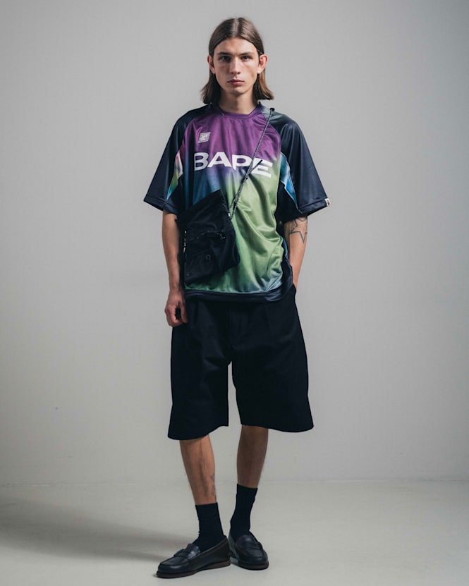 Alt text (EN): Full-body view of a person wearing a multicolor gradient BAPE T-shirt with bold logo print, black wide-leg shorts, black shoes, and a crossbody bag, standing against a plain studio background.