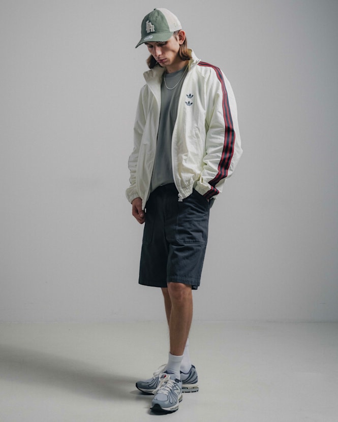 Full-body view of a person wearing a white adidas track jacket with red sleeve stripes, dark shorts, a green cap, and running sneakers, looking down against a plain studio background.