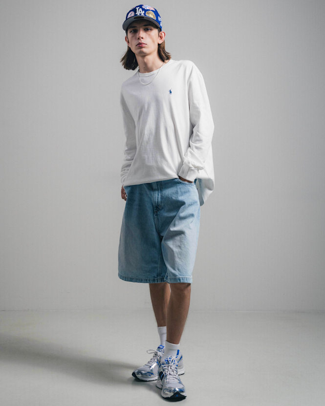 Full-body view of a person wearing a white long-sleeve T-shirt, denim shorts, and blue-accent sneakers, styled with a blue LA Dodgers cap, standing against a minimal light background.