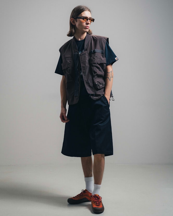 Mid-length view of a person wearing a dark utility vest over a black T-shirt, paired with wide black shorts and orange sneakers, styled with tinted sunglasses against a minimal studio background.