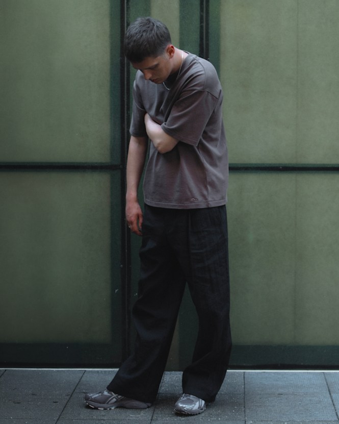 Person wearing a taupe T-shirt, black trousers, and sneakers from the solebox Core Collection, standing in front of glass panels.